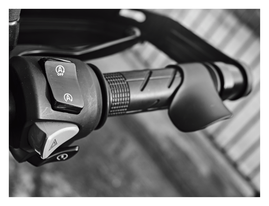 Close-up image of a motorcycle's right handlebar. It focuses on the control cluster, which features a toggle switch for an idling stop system (marked with an "A" inside a circular arrow) and a hazard light button below it. The image also shows the textured throttle grip fitted with a curved plastic palm rest or "cramp buster" attachment. - Google Gemini 3 Pro Preview