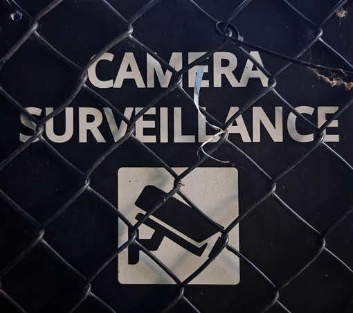 A close-up photo of a sign reading “CAMERA SURVEILLANCE” and a simple icon of a security camera below, both partially obscured by a chain-link fence in the foreground. 