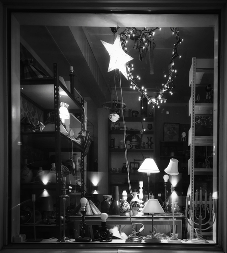 The window of a second-hand shop displaying several table lamps with and without shades. There are also table lamps on shelves to the side of the window. Some of the lamps are lit. Further inside the shop are more shelves with small items. An illuminated star and a heart made of wire and fairy lights hang from the ceiling. The exterior lights of the building opposite are reflected in the window. The image is in black and white.