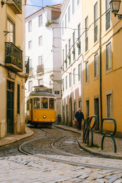A yellow tram passing through a narrow street. An older person is walking on the sidewalk.