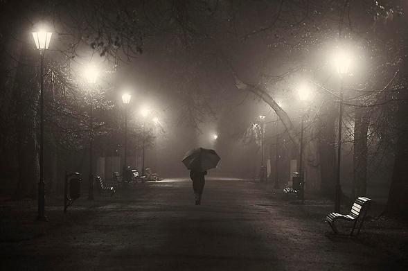 This is a monochrome, sepia-toned photograph of a park promenade at night, shrouded in thick mist. A lone figure holding an umbrella walks away from the viewer down the centre of a wide, paved path. Rows of vintage street lamps on either side illuminate the scene, casting glowing, hazy halos through the fog. The light reflects off the damp ground, creating a soft sheen. Bare, leafless trees line the path, their branches arching overhead into the darkness. Empty wooden park benches are spaced at regular intervals along the edges of the walkway, enhancing the scene's quiet, atmospheric and slightly melancholic mood.