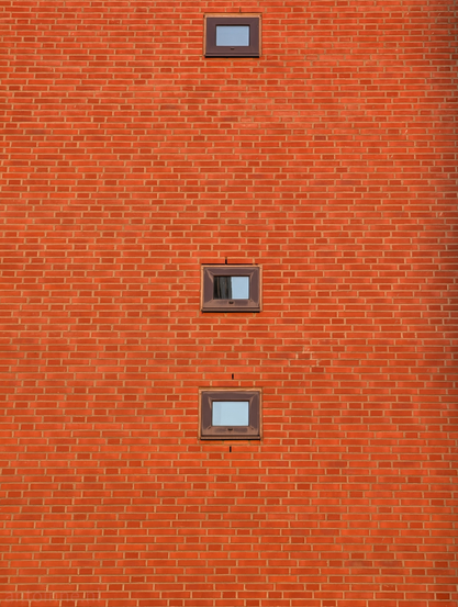 A red brick wall containing three small, vertically aligned windows. 

The wall is constructed of uniform, rectangular red bricks laid in a standard running bond pattern. The mortar lines are light-colored, creating a fine grid texture across the entire frame.

Three identical, square-shaped windows are stacked vertically in the center. They have dark frames that contrast sharply against the warm orange-red of the brick.

The glass in the windows is reflective, showing glimpses of a pale sky and nearby structures, which adds a subtle depth to an otherwise flat, two-dimensional surface.