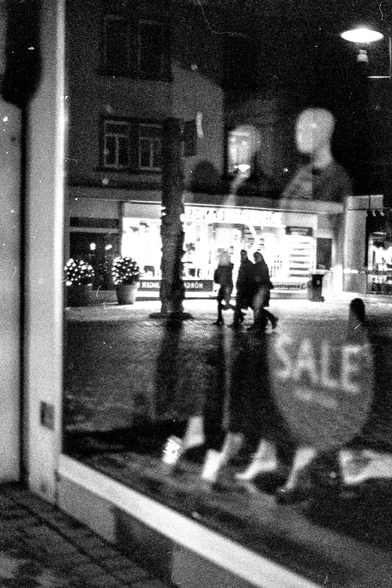 A grainy, high-contrast black and white photograph taken at night, capturing a layered and surreal street scene through a glass window. The image is heavy with film grain and visual noise, giving it a gritty, noir aesthetic.

The composition is split into several visual layers:

    The Foreground (Reflection): A large, bright "SALE" sign is visible in the lower right, appearing as a ghostly reflection. To the right of the center, two featureless mannequins are reflected in the upper portion of the glass, looking like ethereal figures floating over the street.

    The Middle Ground (Street Level): Seen through the glass, a group of three or four people are walking across a dark pavement. They appear as dark silhouettes against a brightly lit storefront in the background. To the left, two decorative, rounded bushes wrapped in fairy lights provide small orbs of glow.

    The Background (Architecture): Above the bright shop windows, the dark outlines of a traditional multi-story buildin