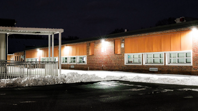 Snowy one-story brick building at night, lit by bright exterior lights with rows of windows along the wall.