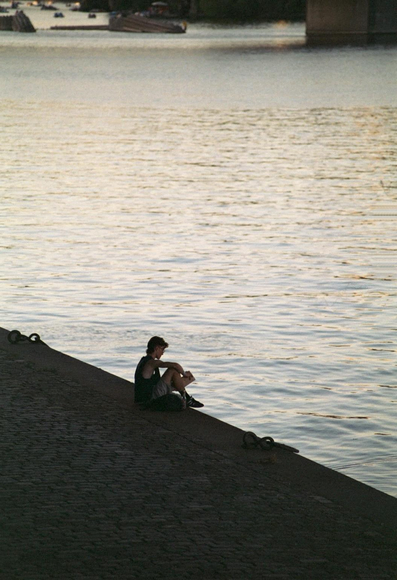 A person sits on a stone embankment beside a river, reading a book. The water reflects the soft light of the setting sun, creating a serene atmosphere. In the background, a few boats are visible on the water.