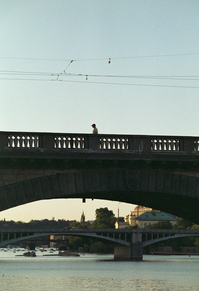 A silhouette of a person walking on a bridge spans across a river. The scene is set during golden hour, with soft light illuminating the surroundings. In the background, another bridge and buildings can be seen along the riverbank.