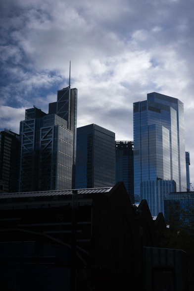 Photo of a cluster of tall shiny buildings some way away, silvered by the daylight from a bright mottled sky. In the foreground at the bottom is a large, relatively low building with an undulating roof, mostly in shadow.