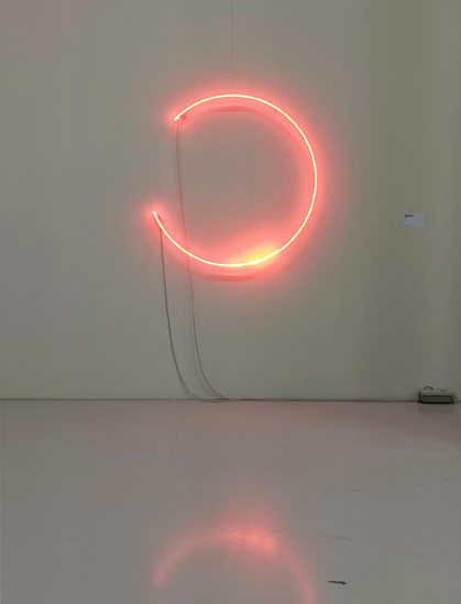 A photo of an orange or pink neon light bent into an incomplete circle mounted on a plain wall, with a power cord running to a power point at the bottom right.