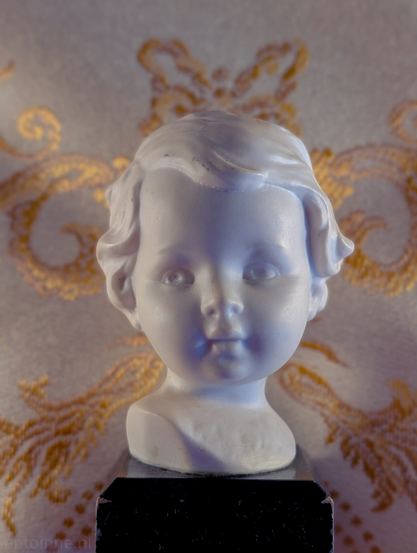 A close-up photograph of a small, white bust of a child. The sculpture is mounted on a dark, block-shaped pedestal.

The sculpture depicts a young child with soft, rounded features. The child has short, wavy hair swept to the side, large eyes, and a small, closed mouth. 

The bust is positioned in front of a textured, light-colored cloth featuring an ornate golden-yellow floral pattern. The warm colors of the background provide a sharp contrast to the cool lighting on the statue.
