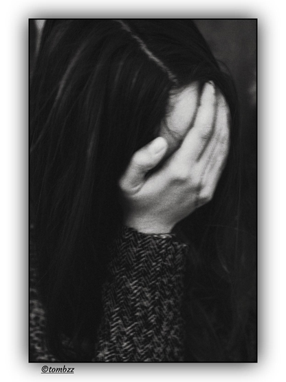 The black and white analog photograph shows a young woman with long, dark and straight hair covering her face with her hand. Her features remain hidden, making it a faceless portrait filled with ambiguity. Soft light and a subtle vignette give the image an intimate, quiet character. The gesture of covering her face can suggest shame, defiance, protection, or a moment of inward focus… it stays open to interpretation. The frame balances presence and absence, creating space for emotions that remain unnamed.