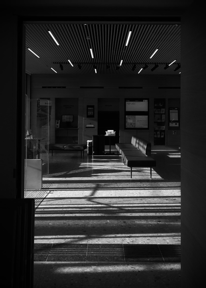 das bild zeigt den blick durch eine türöffnung in einen öffentlichen raum, eine wartehalle oder eine bibliothek. das bild ist in schwarzweiß gehalten, uns es sind keine menschen zu sehen, was eine gewisse nüchternheit und formalität unterstreicht.
das auffälligste merkmal sind die starken licht- und schattenmuster; zum einen durch die horizontalen lamellen und lichter an der decke; zum anderen durch das von links einfallende licht. diese muster erstrecken sich über den boden und die darauf befindlichen sitzgelegenheiten, was eine interessante grafische wirkung erzeugt.
im hintergrund sind informationstafeln und schalter zu erkennen, was den öffentlichen charakter des ortes weiter betont. die komposition ist sehr aufgeräumt und geometrisch, was dem bild eine gewisse strenge verleiht.