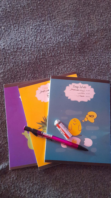Three school notebooks with colored covers in purple, yellow, and blue, with a pencil and eraser, resting on a textured gray surface. The blue notebook features a cartoon illustration of a chick saying "NO!" and the text "Easy Write" in English and Arabic.