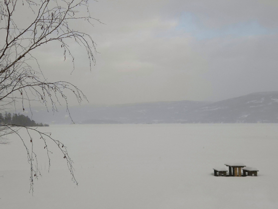 A frozen and snow-covered lake, with snow falling only on the other side, giving the hills behind a soft shape and pastel white colour. In the foreground is a snow-covered picnic table with two benches. It is cloudy, but you can glimpse some blue sky.