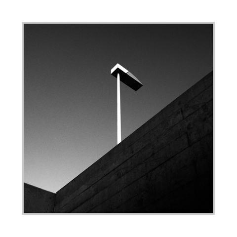 Square black-and-white photograph. The image is a low-angle view of a streetlamp rising from behind a concrete wall: a slender bright pole stands near the center, topped by a rectangular lamp head with a sharply angled edge. A large concrete surface cuts diagonally across the lower half from left to right, showing horizontal formwork lines and darker shading toward the bottom right, with a second wall corner visible at the lower left. The sky fills the upper area with a smooth gradient from lighter gray near the horizon to darker gray at the top.