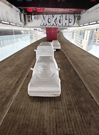 An upside down photo of three life-size car models made from white paper or plastic suspended below a concrete bridge with city buildings visible below. By putting the photo upside down the white cars seem to drive on a concrete surface.