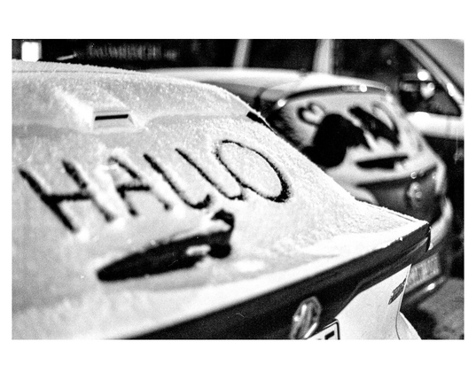 A written "Hallo" into snow covering the rear window of a car. Another car behind shows a  heart-sign on its snow-covered rear window. Black-and-white photography.