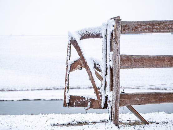 Ein Flügel eines Ranchzaunes ist nach innen geöffnet, das Holz ist schneebedeckt.
Das Feld, der Hang im Hintergrund weiß von Schnee. Sehr heller Himmel.