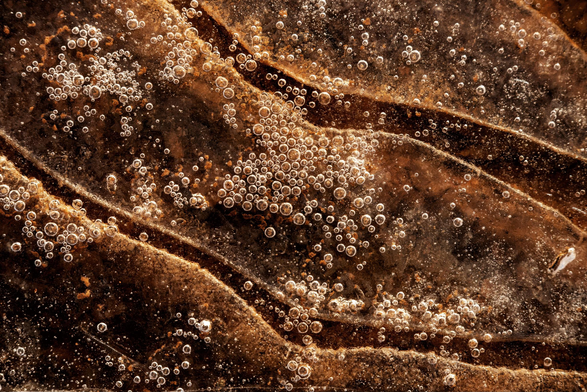 Das Bild ist eine Nahaufnahme einer gefrorenen Oberfläche die durchscheinend ist und eine darunter liegende, dunkelbraune, fast rostfarbene Textur zeigt. Die Oberfläche ist uneben und weist mehrere geschwungene, wellenartige Erhebungen oder Rillen auf, die diagonal von links unten nach rechts oben verlaufen. Über die gesamte Fläche sind unzählige kleine, runde Luftblasen verteilt, die im Gegenlicht hell leuchten und in Gruppen angeordnet sind, insbesondere in der Mitte des Bildes. Die Beleuchtung betont die Textur und die Transparenz des Eises und erzeugt starke Kontraste zwischen den glänzenden Blasen und dem dunklen, strukturierten Hintergrund. Die Farben sind überwiegend warme Brauntöne, Gold und Weiß.