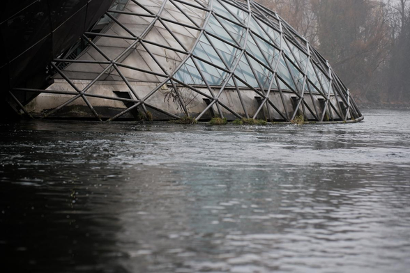 Im Vordergrund bis zur Mitte ist Wasser. Dahinter sieht man einen Teil eines futuristischen Gebäudes, das eine Insel auf dem Wasser darstellt. Es hat außen diagonale Verstrebungen und darunter Metall und Glas.