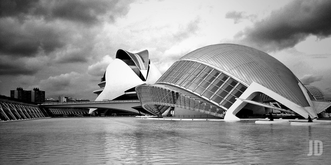 Esta imagen captura la espectacular arquitectura modernista de la Ciudad de las Artes y las Ciencias en Valencia, España, diseñada principalmente por Santiago Calatrava.
Una toma en blanco y negro con alto contraste que acentúa las formas futuristas y orgánicas de los edificios.
En primer plano se aprecia L'Hemisfèric (con forma de ojo), y al fondo destaca la silueta del Palacio de las Artes Reina Sofía.
Los edificios parecen "flotar" sobre grandes estanques de agua cristalina que reflejan sus estructuras, bajo un cielo dramático lleno de nubes densas.
El uso del monocromo resalta la textura del hormigón blanco y las superficies acristaladas, dándole un aire atemporal y casi de ciencia ficción.