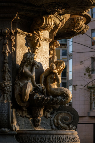 Español: detalle de una fuente de piedra ornamentada con figuras escultóricas de estilo clásico, iluminadas por luz cálida, con edificios urbanos desenfocados al fondo.

English: close-up of an ornate stone fountain featuring classical sculpted figures, bathed in warm light, with blurred urban buildings in the background.