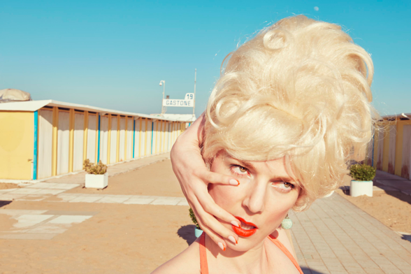 sur les planches au bord de la mer, entre deux rangées de cabines de bain, une femme blonde à la coiffure élaborée passe son bras gauche derrière sa tête pour poser son annulaire dubitatif sur la commissure droite de sa bouche rouge