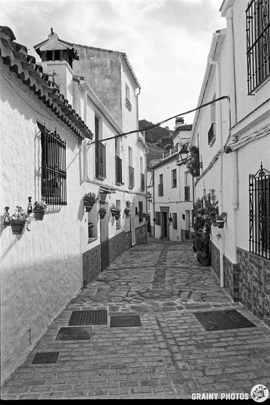 A narrow, cobblestone alleyway lined with whitewashed buildings, decorative wrought-iron details, and potted plants, leading to a serene, mountainous backdrop. The scene captures a peaceful, historic atmosphere in black and white.