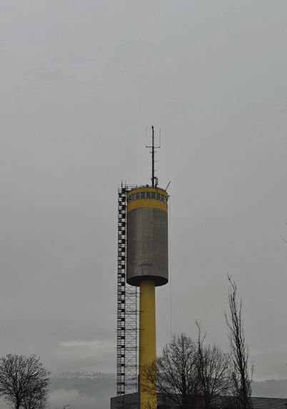 A photo of a tall tower consistng of a round metal cylinder with a yellow band, windows and antennas placed on top of a tall yellow pillar. It stand  in front of a gray sky, scaffolding is running down the side and bare trees can be seen at the bottom.