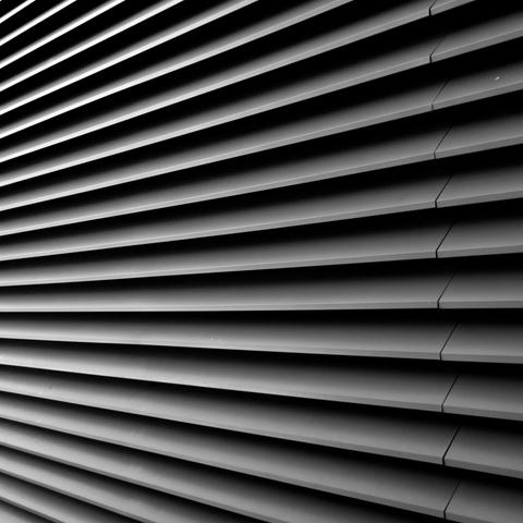 Monochrome photo of a wall of slim metal slats taken from side on so they look like they're radiating out from a point somewhere to the left of the frame. Towards the right edge, they each have a small discontinuity, suggesting that this is a cover for a door.