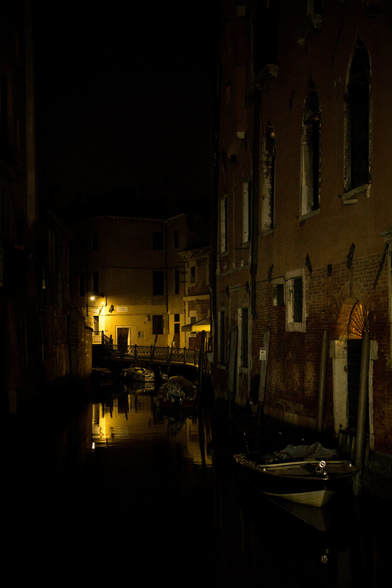 In the dark a canal slips by old brick buildings. A yellow light on a white wall shades a doorway and glints from the water's surface, the boats that float upon it buttoned up against the night.