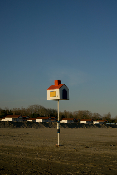 On a pole on a beach sits a small house with white walls and a red roof. Like an angler fish's lure, it attracts larger buildings that find themselves trapped behind a wall of sand. What it wants with them, no-one knows. For now they stand together under a denim blue sky.