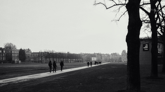 black & white winter scene of a large open space with a footpath cutting across diagonally. dark silhouette’s of people walking are on the path. there are two barren trees at the right foreground, and between them is a sign on a building that reads “Van Gogh Museum”. four storey buildings are visible in the far background, behind a row of barren trees. the sky appears bright and cloudless. 