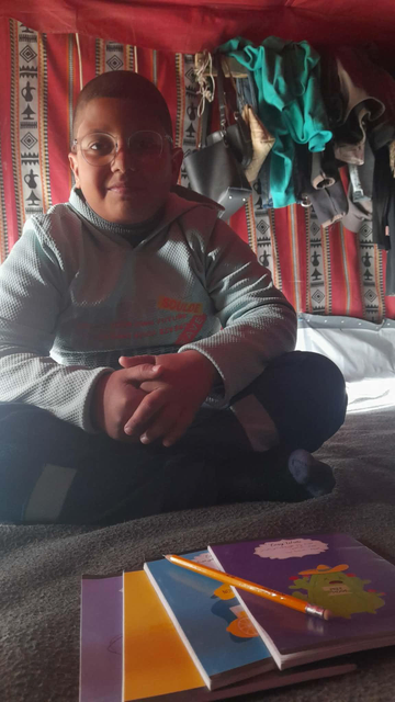 A young boy (Elias) with glasses wearing a light blue sweatshirt sits cross-legged on a dark blanket with a stack of notebooks and a pencil. Red and white patterned fabric hangs in the background.