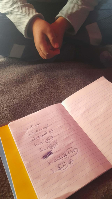 Open notebook on a lap with English words and Arabic script, child's hands clasped in the background.