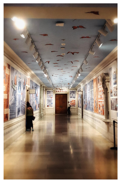 A view of a hallway inside the New York Public Library (at 42nd Street), with the walls at the sides receding towards a wooden door at the center. Illustrated monarch butterflies are displayed on the ceiling, giving the impression of a butterfly migration in a pale blue sky. On the walls are oversized insect-themed illustrations in comic book-style panels, illuminated by ceiling-mounted spotlights. A couple of library patrons are half-hidden by a doorway to the left. A white-shirted security guard stands by the door at the far end. The wide marble-tiled floor gives off a reflective sheen.