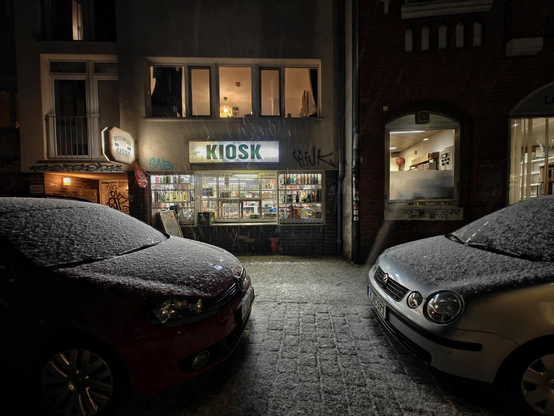 A photo of a small illuminated kiosk storefront at night, flanked by two snow-covered parked cars.