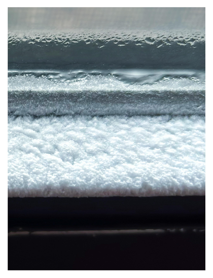 A minimalistic image of horizontal bands of different widths and textures, in varying light and dark tones. It’s actually an extreme closeup of a layer of white frost on the lower lip of an interior window sash, with some drops of condensation on the glass above it and a blurred glimpse of the wire mesh screen on the outside.
