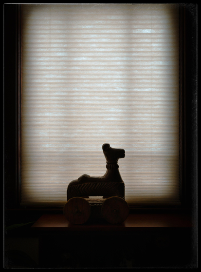 A silhouette of a dark stained, 10 inch, carved wooden toy sitting on a shelf in front of a cellular beige window shade. The simple carved horse has large, working wheels that rise up to just below the horse's belly.
