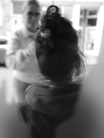 Das Schwarzweißfoto zeigt 2 Frauen, durch die Scheibe eines Cafés fotografiert. Die Frau im Vordergrund wendet dem Betrachter den Rücken zu und ist durch Bewegung leicht unscharf abgebildet. Die Frau im Hintergrund ist dem Betrachter und der anderen Frau zugewandt. Sie ist nicht im Fokus und dadurch nur undeutlich zu erkennen. Sie scheint zu lächeln. Im Hintergrund scheint eine Straße zu sein: ein Auto und eine Hausfassade sind schemenhaft sichtbar.

The monochrome photograph depicts two women, photographed through the window of a café. The woman in the foreground has her back to the viewer and is slightly out of focus due to movement. The woman in the background is facing the viewer and the other woman. She is not in focus and therefore only vaguely recognizable. She appears to be smiling. There appears to be a street in the background: a car and a house facade are vaguely visible.