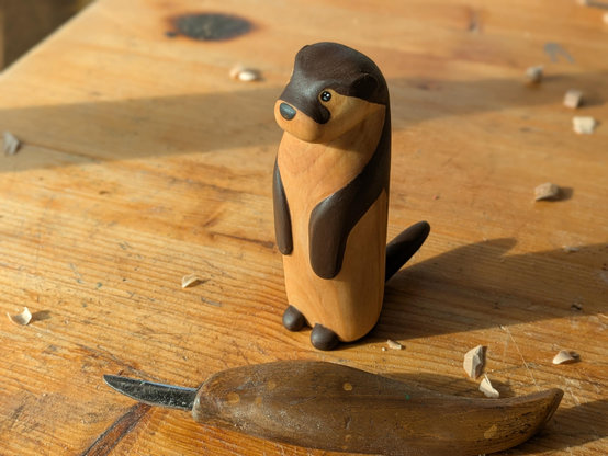 Woodcarved standing up otter on a workbench, next to a carving knife and a few wood chips.