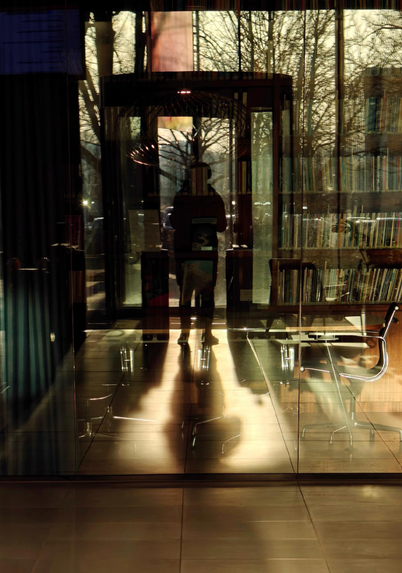 A photo showing a silhouetted person, standing in front of a revolving glass door, reflected in a glass wall, with bookshelves, chairs, and trees outside layered through overlapping reflections.