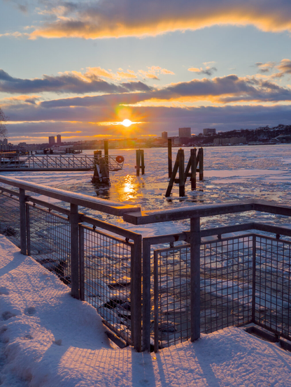 The sun was about to set over a mostly frozen Hudson River, with some clouds in the sky. In the foreground, the golden sun cast shadows of the metal fence on the deep snow.
