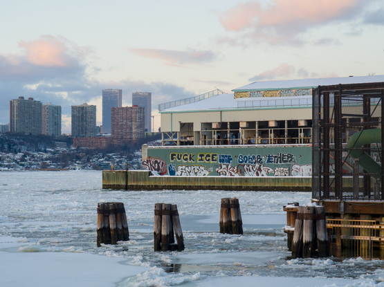 A few wooden pillars stick out of a mostly frozen Hudson River. In the distance is an industrial building with some graffiti, one of which says Fuck ICE.