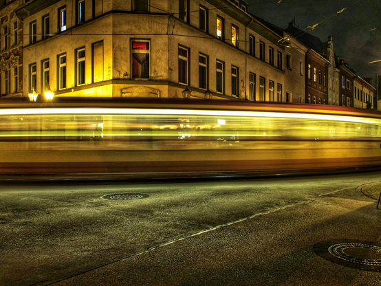 Die Szene zeigt eine nächtliche Straßenkreuzung in einer Stadt, dominiert von einem starken Bewegungseffekt. Im Vordergrund befindet sich eine asphaltierte Straße mit zwei runden Kanaldeckeln. Unmittelbar darüber, horizontal durch das Bild verlaufend, erstreckt sich ein langer, verschwommener Schweif aus hellem, gelbem Licht, der die vorbeifahrende Straßenbahn aufgrund einer Langzeitbelichtungseinstellung darstellt. Dieser Lichtstreifen ist das zentrale Motiv. Im Hintergrund steht ein mehrstöckiges, älteres Gebäude im Eckbereich, dessen Fassade gelb beleuchtet ist. Die Fenster des Gebäudes sind ebenfalls hell erleuchtet, was auf bewohnte Räume hinweist. Die Architektur ist typisch für eine europäische Altstadt.