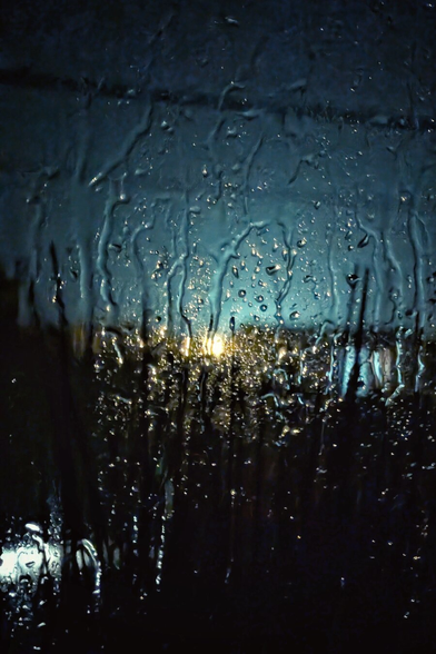 A vertical photograph taken looking out through a glass window at night. The pane is drenched in water, with heavy streaks of rain and large droplets distorting the view. Through the wet glass, the outside world is a wash of deep midnight blues and blacks, punctuated by blurred, glowing yellow and orange lights from distant streetlamps or buildings. The image is moody and atmospheric, emphasising the cold weather outside compared to the implied warmth of the viewer's indoor perspective.