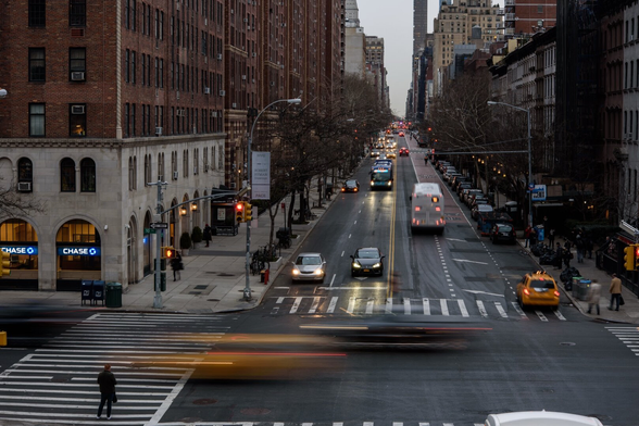 A busy street scene in the city with cars, including a yellow taxi, moving along the road. Tall buildings can be seen in the background, and a Chase Bank sign is visible. The streetlights are lit, and there are a few pedestrians on the pavements. The atmosphere is lively, partly due to motion blur.