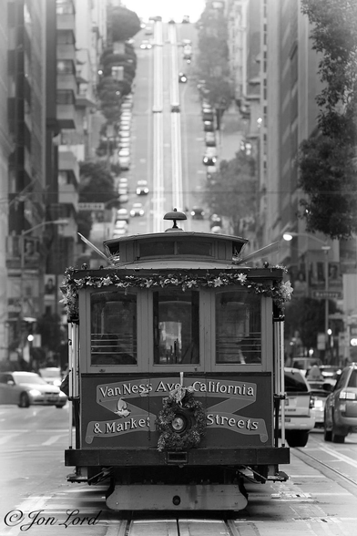 This is a black and white street photo in portrait format of a stationary San Francisco cable car (2012).

The camera is directed along a straight and wide city street. Below the centre of the image is an iconic San Francisco cable car, stationary, in the middle of the street and at the start of the line.  The cable car car is viewed from the front (or perhaps the rear?) (When it gets to the end of the line the driver gets out of the front goes to the rear and doubles back in reverse, though on a parallel track). The cable car is all wood above the metal chassis. On the lower part of the front/back are the words - 'Van Ness Ave California & Market Streets'. Above, are three rectangular windows with a slightly arched roof above. It's a month before Christmas and there is tinsel around the edge of the roof. On either side of the cable car is the usual assortment of city traffic. On either side of the street are the bottom few floors of various high-rise buildings. In the distance, about half a km, the street climbs steeply with the summit being just short of the top of the image, a little strip of white sky is visible in that gap. Where the road climbs so steeply, the two parallel sets of tracks can be seen clearly in the middle of the street. 

The location: California Street at the junction with Market Street, looking west, in San Francisco's Business district. 