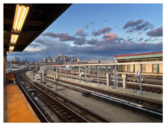 At an elevated outdoor subway station in Brooklyn. Train tracks in the foreground recede towards the left of the image, then curve right towards the horizon. At left is the edge of the covered platform: a row of fluorescent lights above, a broad yellow safety strip below. Far off, the skyline of lower Manhattan stands under a sky at sunset: deepening blue, with masses of dark clouds edged in gold.