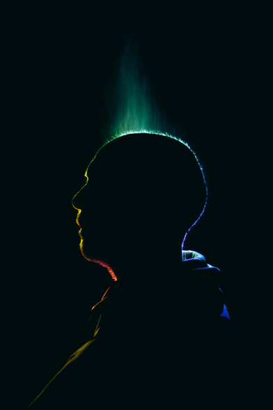 A photograph of a silhouette of a person's (Peter) profile against a black background, illuminated by a colourful light gradient. The gradient starts with green at the top, transitioning through yellow and red to blue at the bottom. The light creates a neon outline around the contours of his head and shoulders, emphasizing his profile.