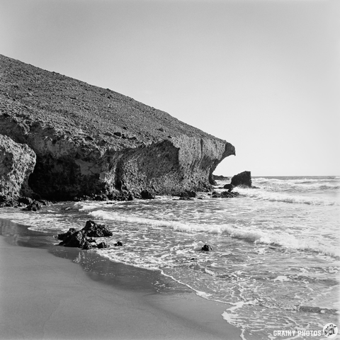 A black and white image of a rocky beach with gentle waves lapping at the shore. A steep cliff rises on one side, contrasting with the calm sea, creating a serene coastal landscape.